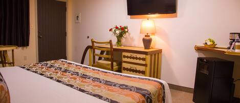A hotel room with a bed, a desk, a chair, a television, and a small refrigerator.