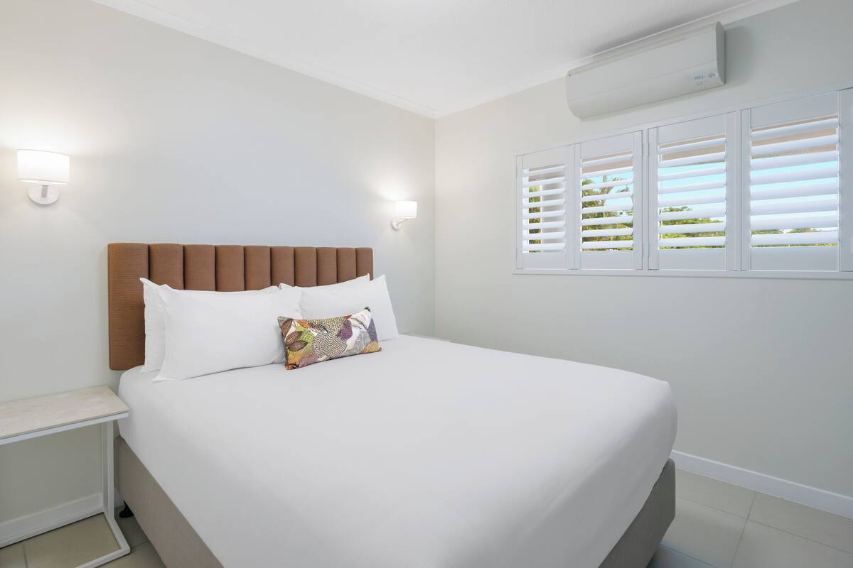 A neatly made bed with white linens and a brown headboard, a bedside table, and a window with white blinds.