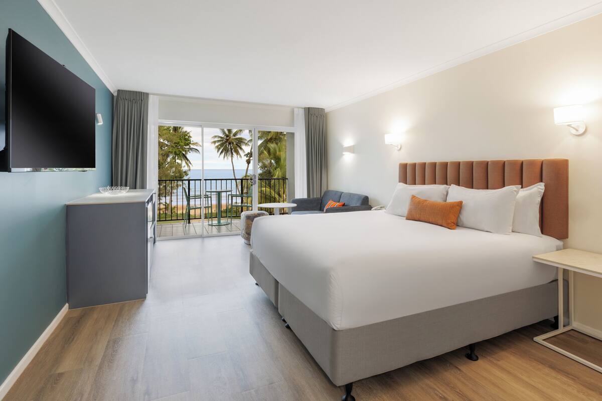 A hotel room with a large bed, a TV mounted on the wall, a small table, and a balcony with a view of palm trees and the ocean.