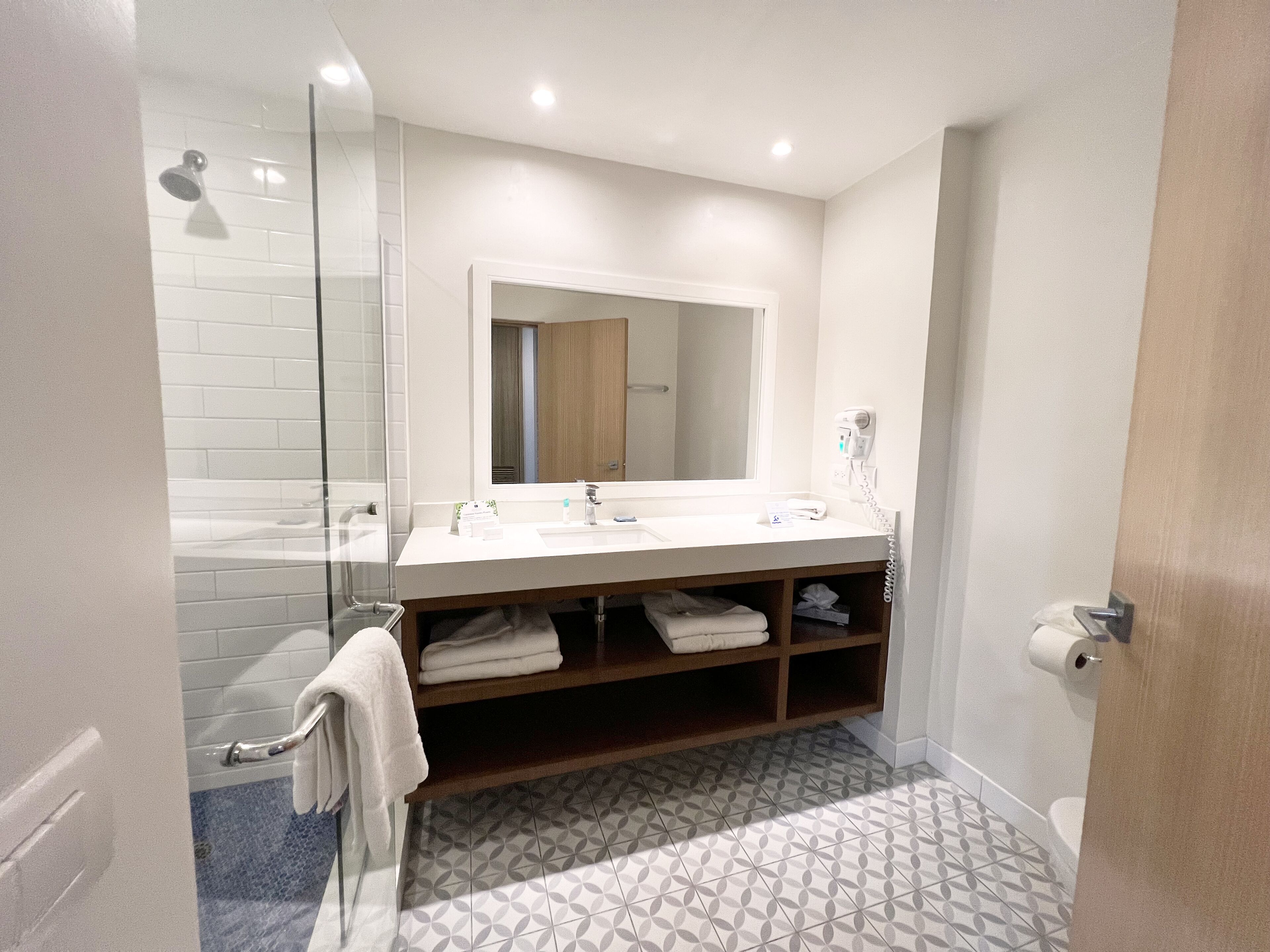A modern bathroom with a glass shower enclosure, a white sink with a mirror above, and patterned floor tiles.