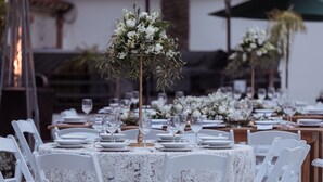Un montaje formal para comidas al aire libre con sillas y mesas blancas, cada juego con copas de vino y platos, y decorado con centros de mesa florales.