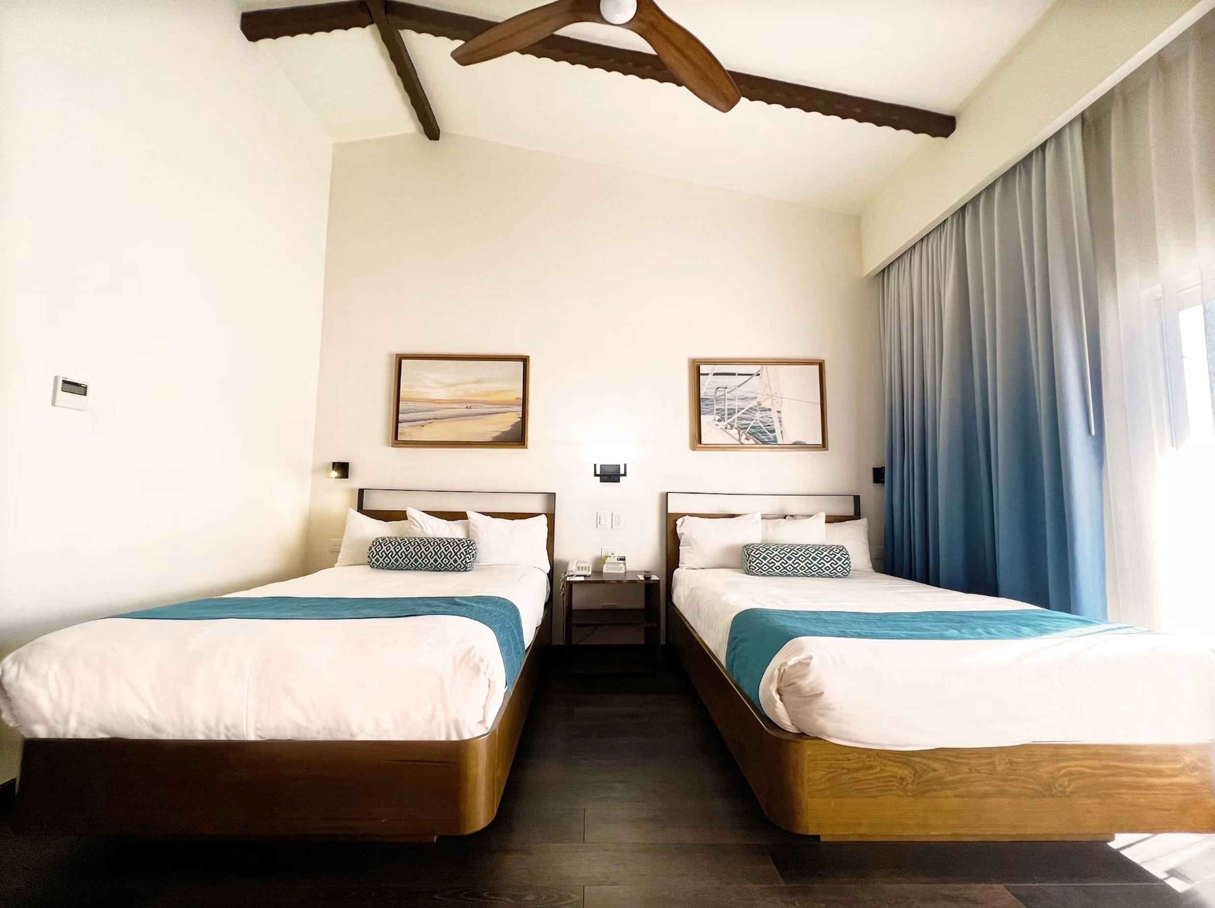 A hotel room with two single beds, a ceiling fan, a nightstand with a lamp, and artwork on the walls.