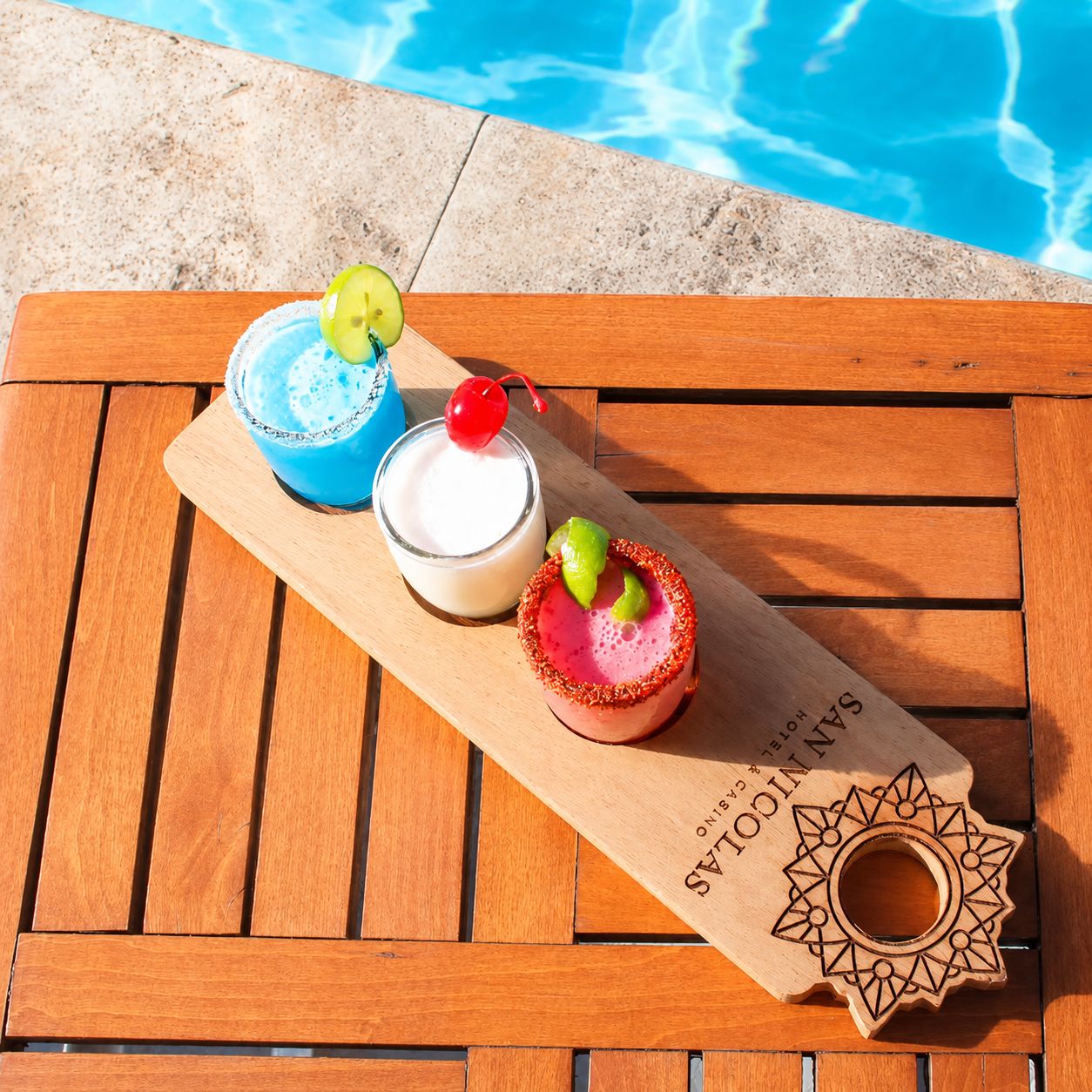 Una bandeja de madera con dos bebidas y un adorno junto a una piscina.