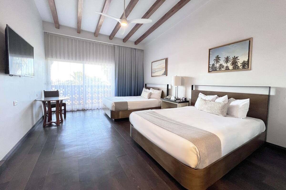 Een hotelkamer met twee bedden, een bureau, een stoel, een plafondventilator en een groot raam met gordijnen.