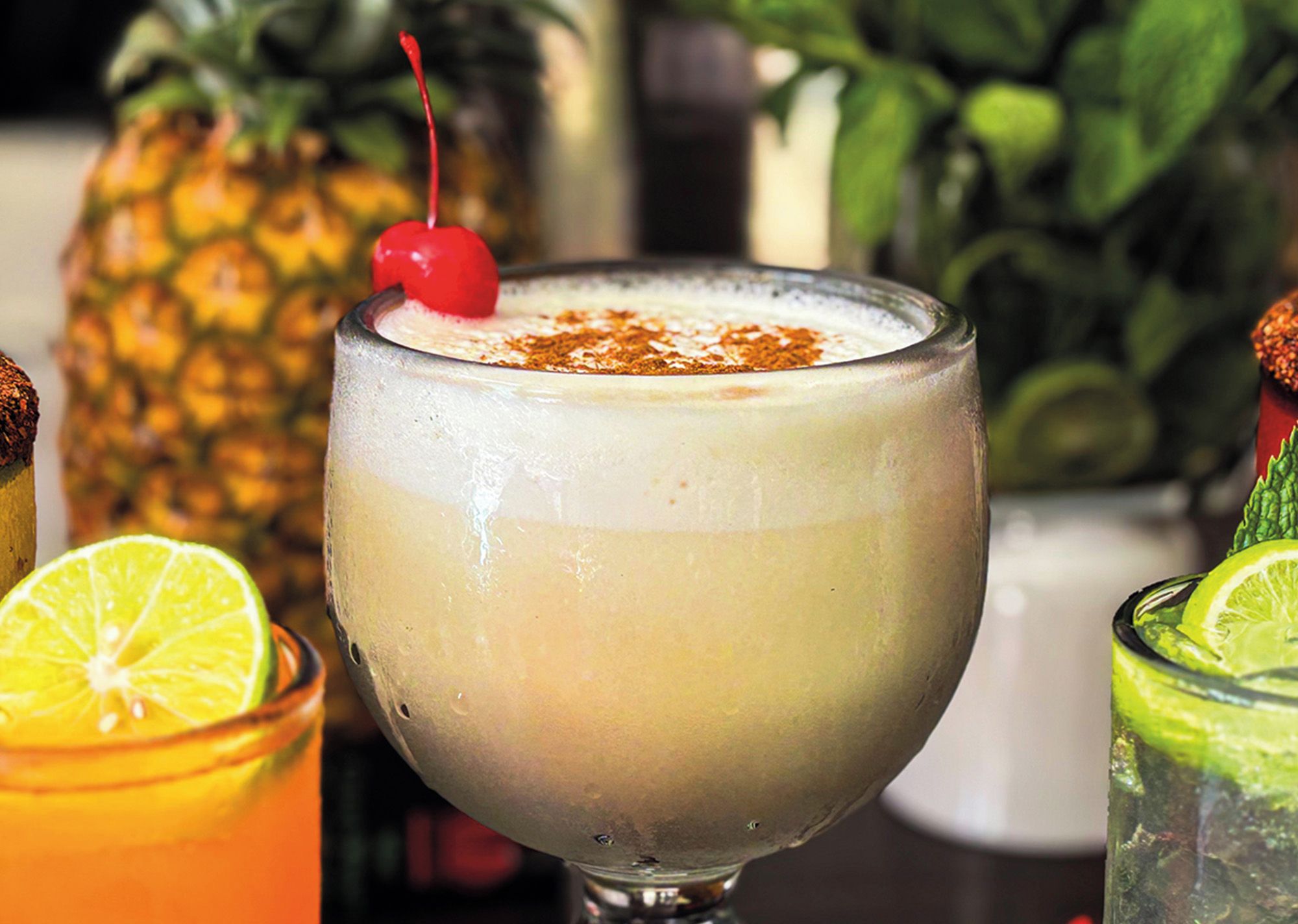 A cocktail in a glass with a cherry on top, surrounded by other drinks and a pineapple.