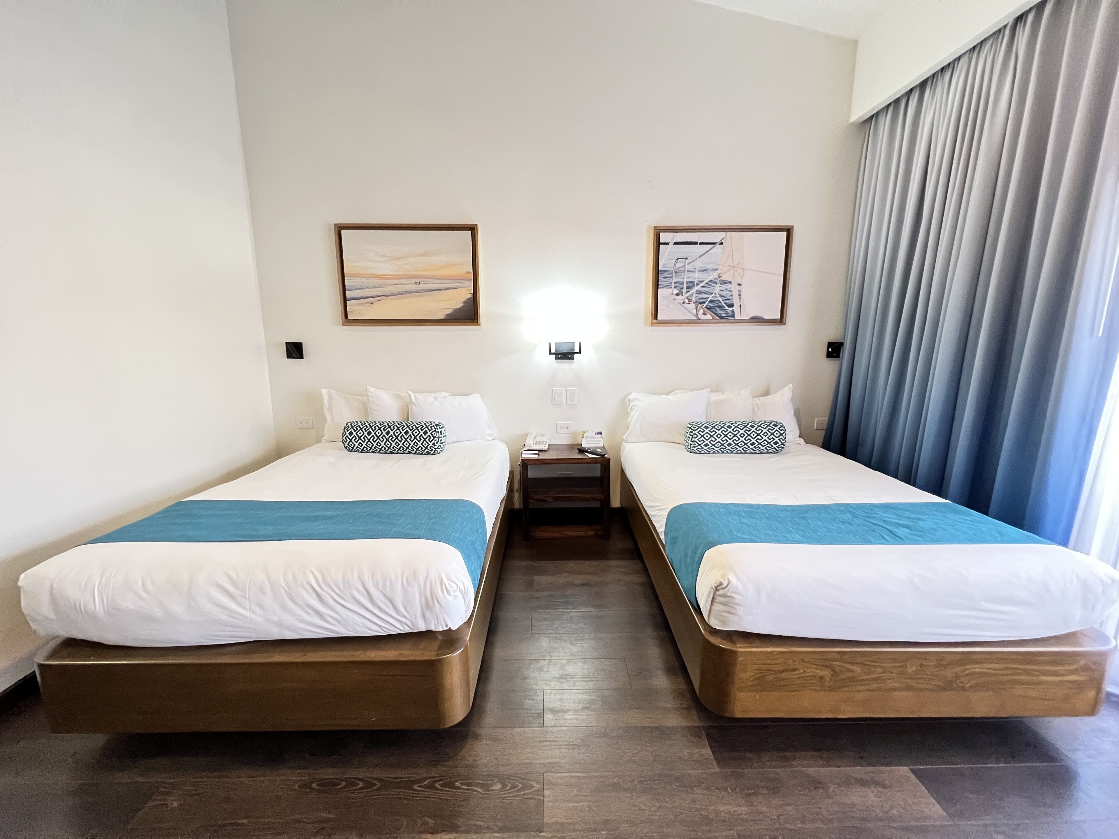 A hotel room with two single beds, a nightstand with a lamp, and two framed pictures on the wall.