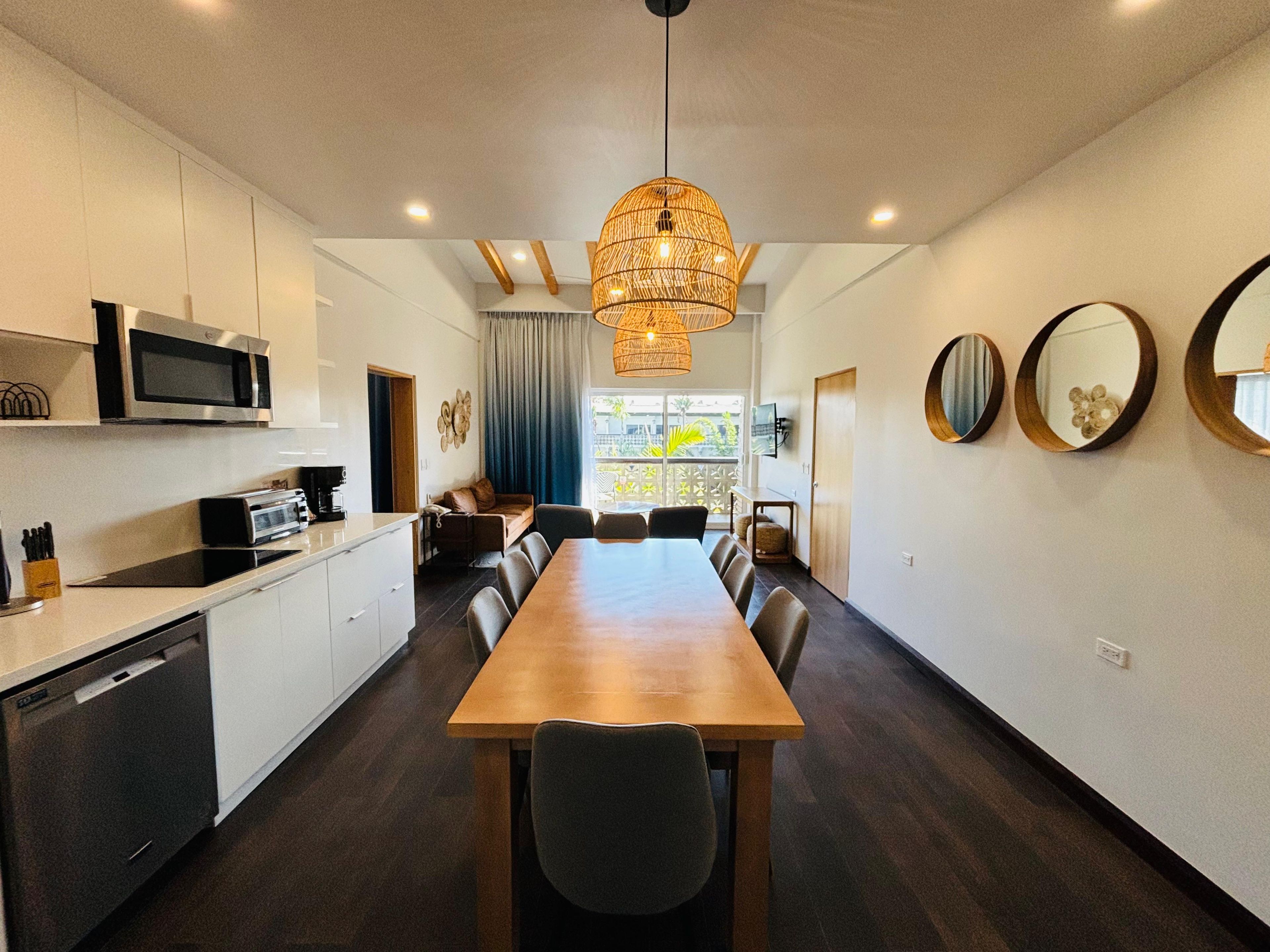 A modern kitchen with a dining area, a large wooden table, and a hanging light fixture.