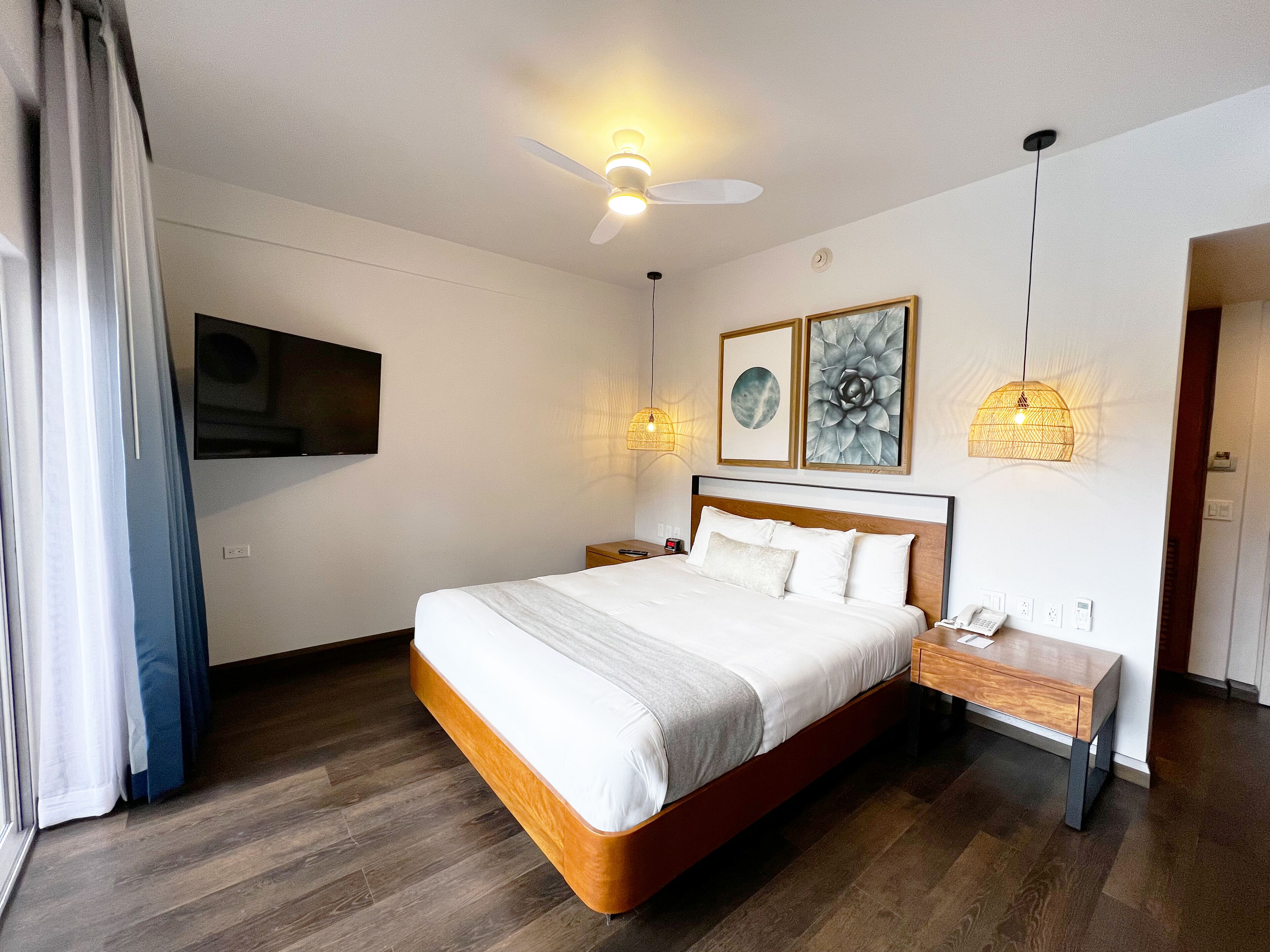 Una habitación de hotel moderna con una cama grande, un ventilador de techo, un televisor montado y obras de arte en las paredes.