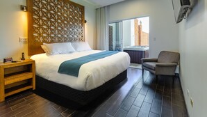 Habitación de hotel con una cama grande, una mesita de noche de madera, una silla y vistas a un edificio a través de una puerta de cristal.