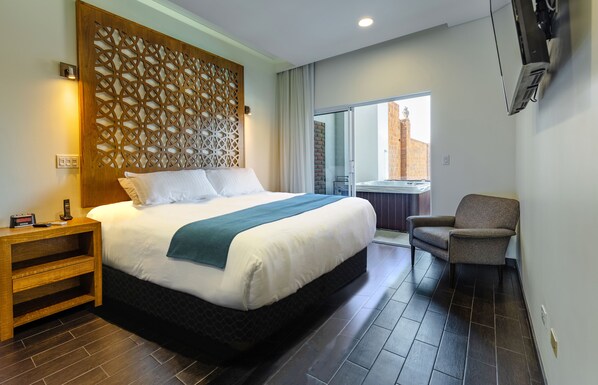 Habitación de hotel con una cama grande, una mesita de noche de madera, una silla y vistas a un edificio a través de una puerta de cristal.