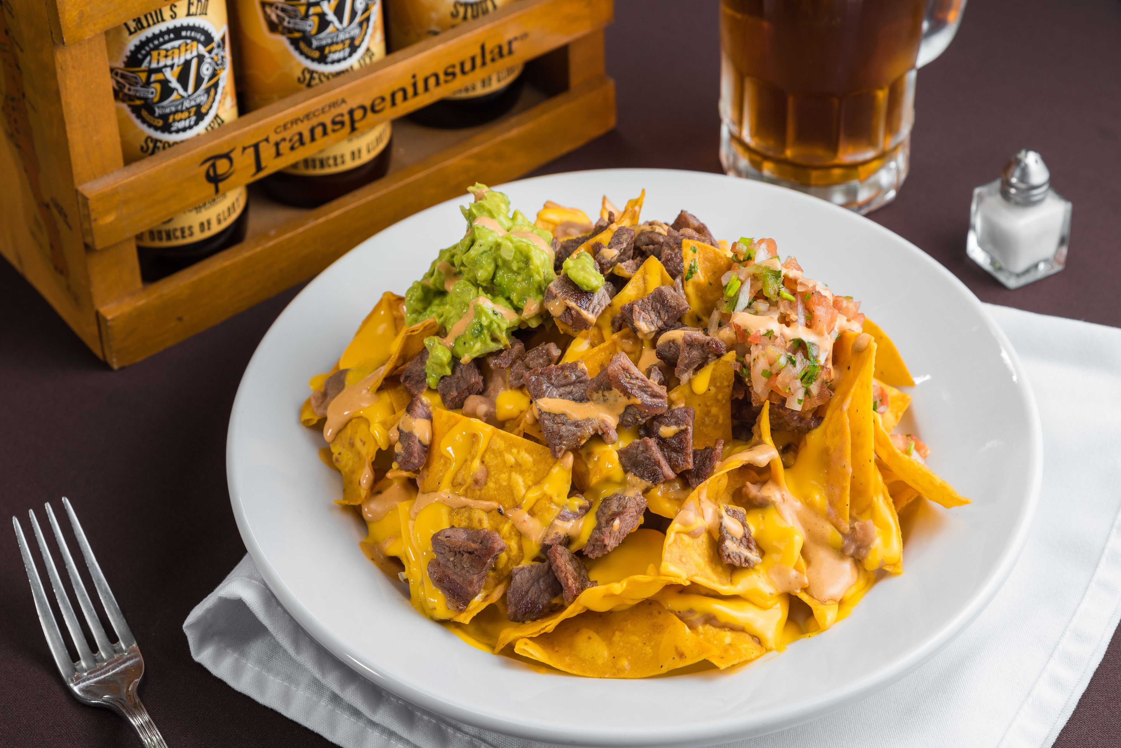 Une assiette de nachos garnis, un salier et une caisse de bières en arrière-plan.