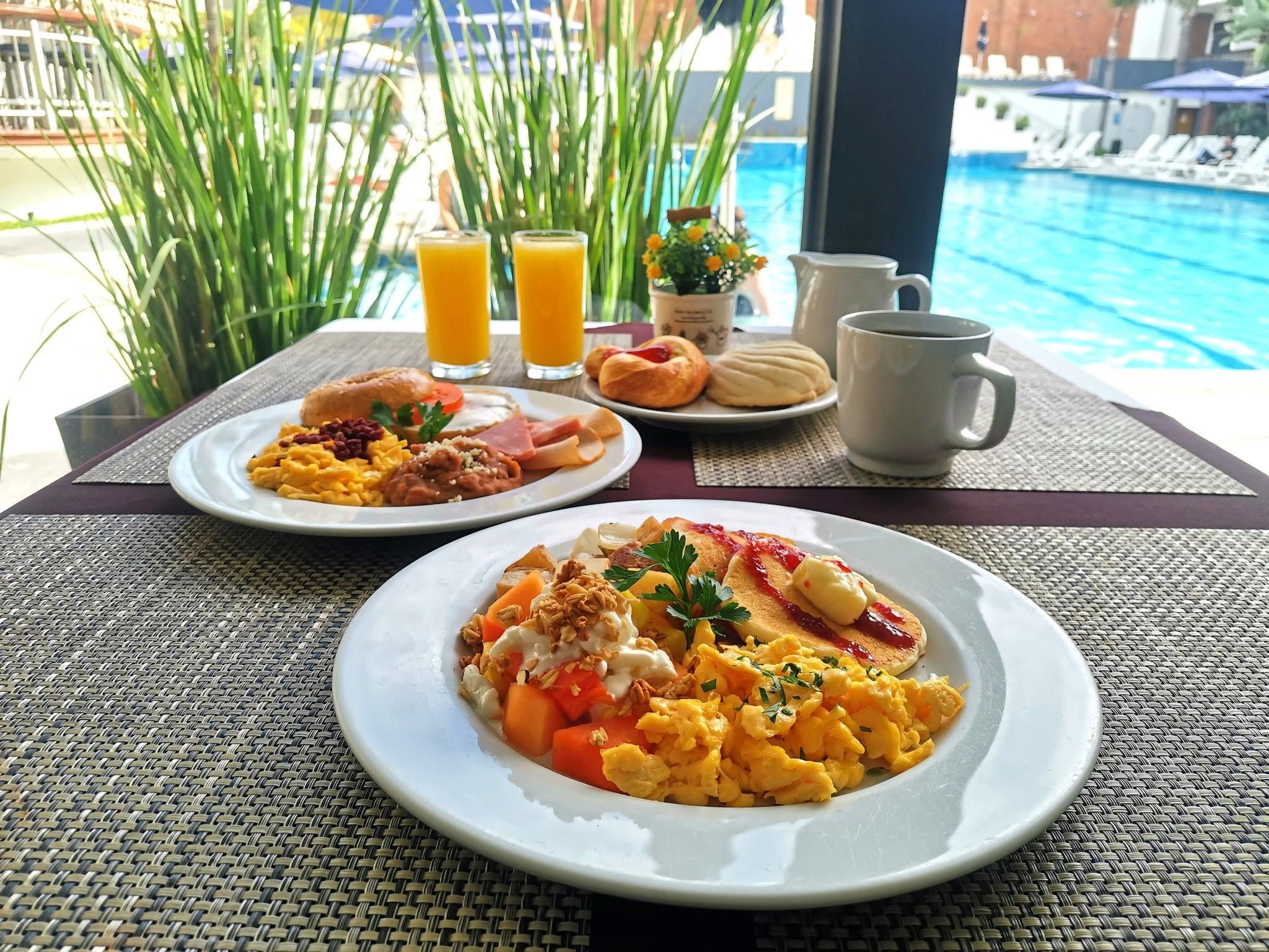 Two plates of breakfast with eggs, ham, and toast, a pitcher of coffee, and two glasses of orange juice on a table by a pool.