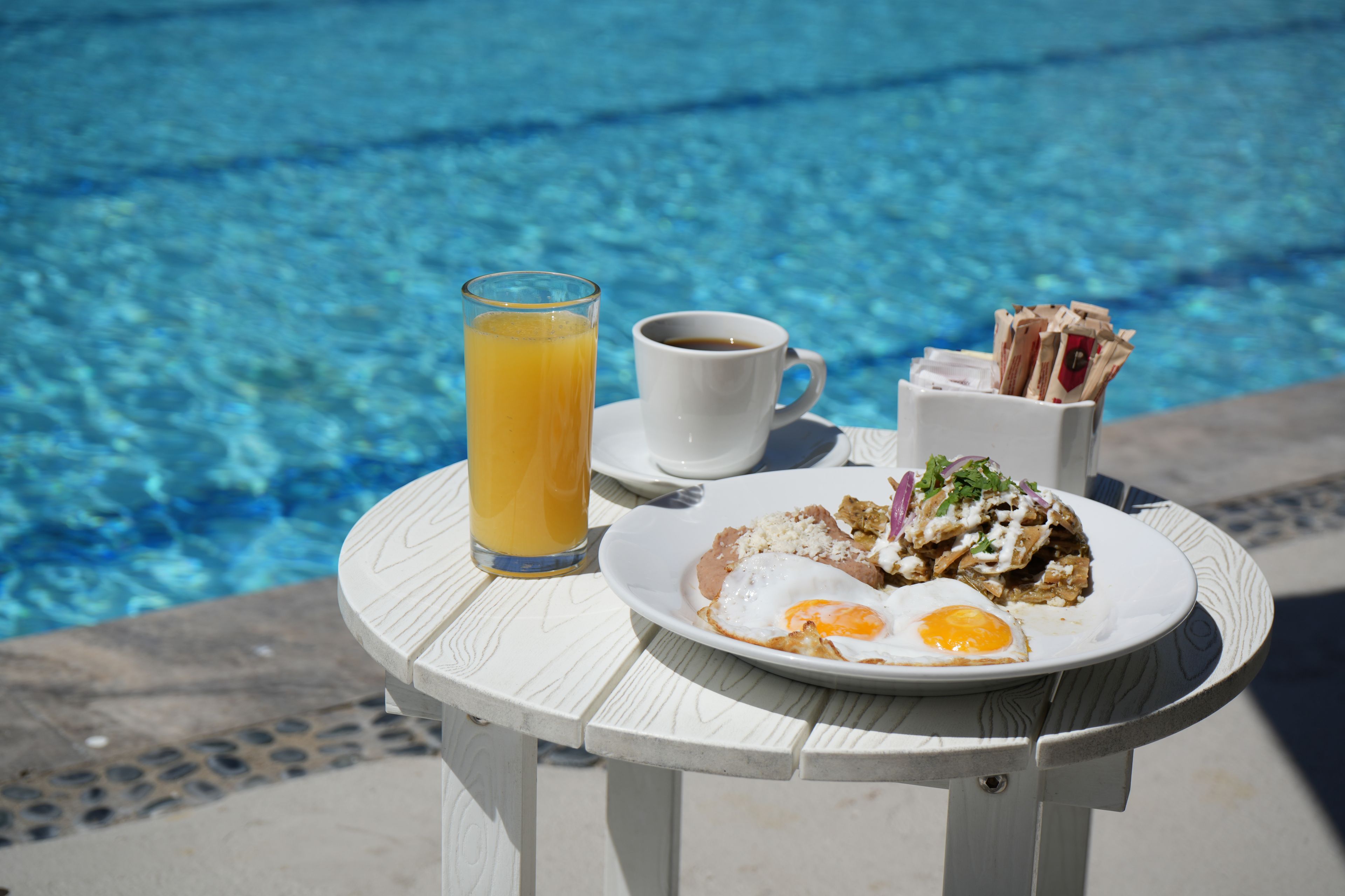 A table with a plate of food, a glass of orange juice, and a cup of coffee by a pool.