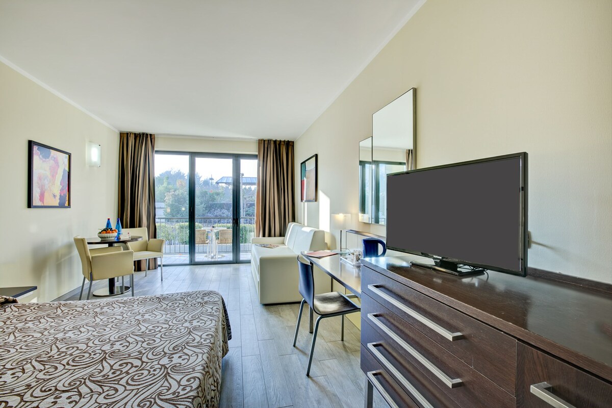 A hotel room with a bed, a desk, a television, and a balcony with curtains.