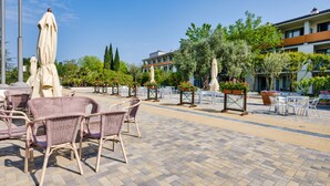 A paved outdoor area with wicker chairs and tables, surrounded by potted plants and umbrellas. - Parc Hotel (Castelnuovo del Garda)