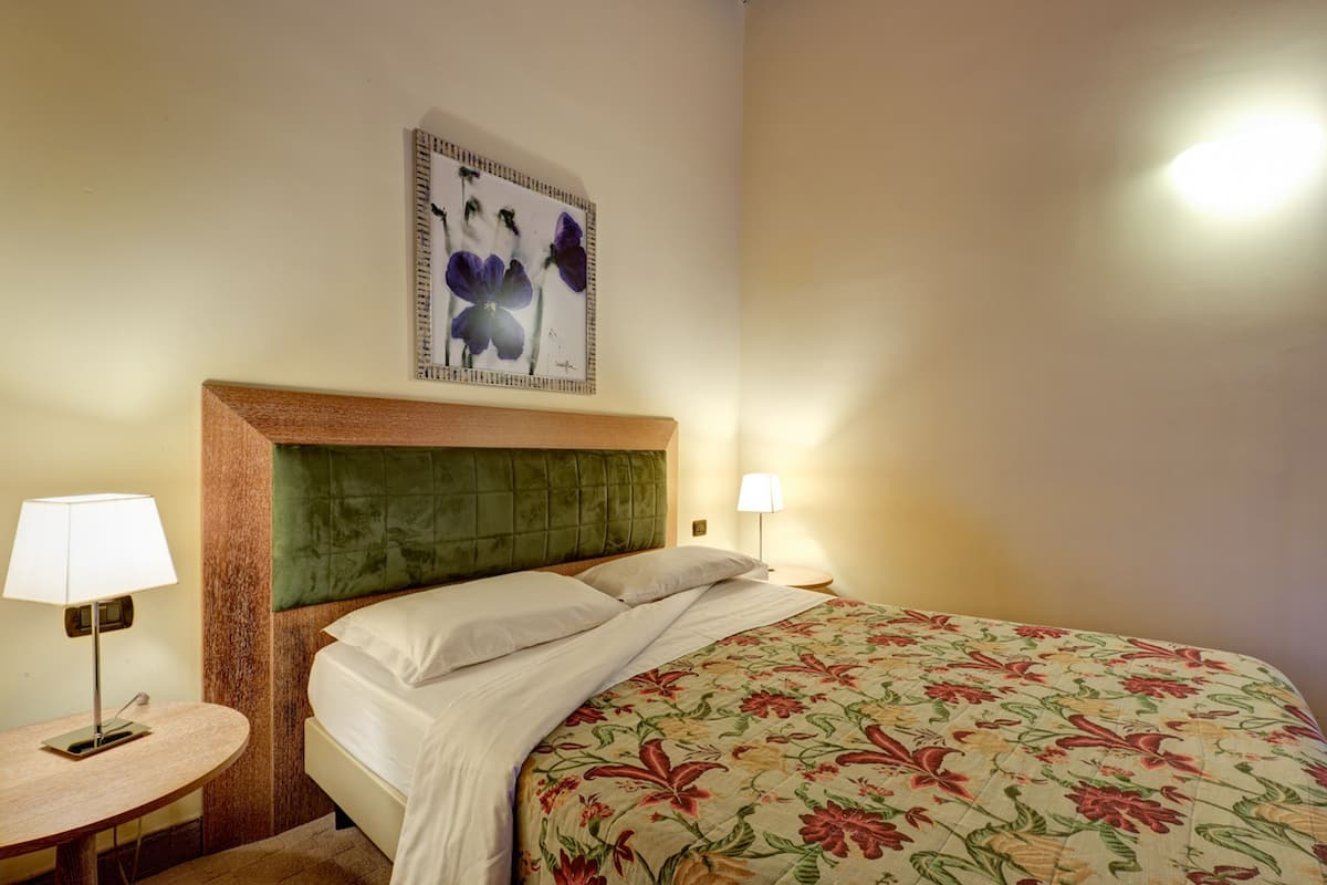 A hotel room with a bed, a nightstand, a lamp, and a framed floral artwork on the wall.