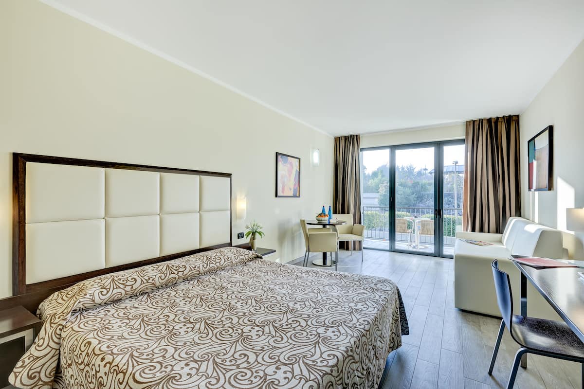 A hotel room with a large bed, a desk, a chair, a sofa, and a balcony with a view.