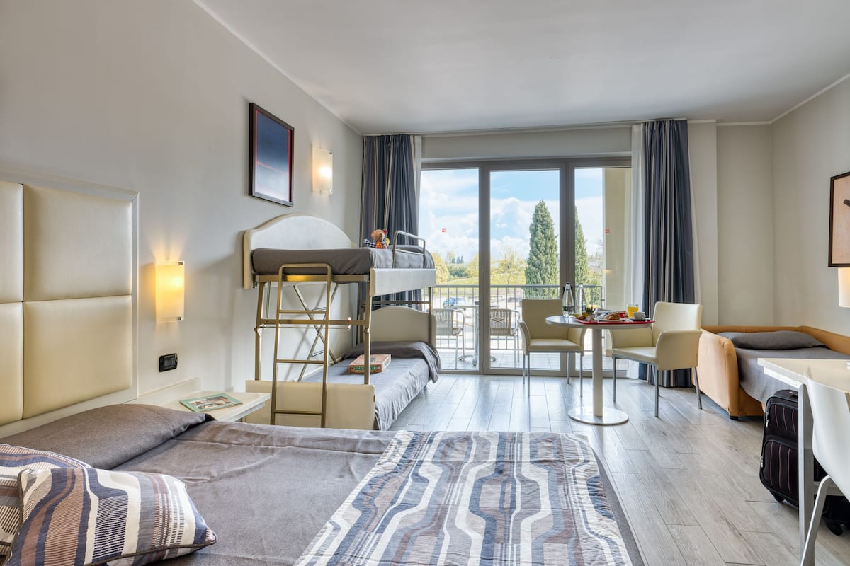 A hotel room with a bunk bed, a desk with a chair, a small table, and a view of the outdoors.
