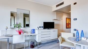 Une chambre d’hôtel moderne avec un bureau blanc, un téléviseur à écran plat, une commode blanche, une chaise, une petite table et une poubelle.