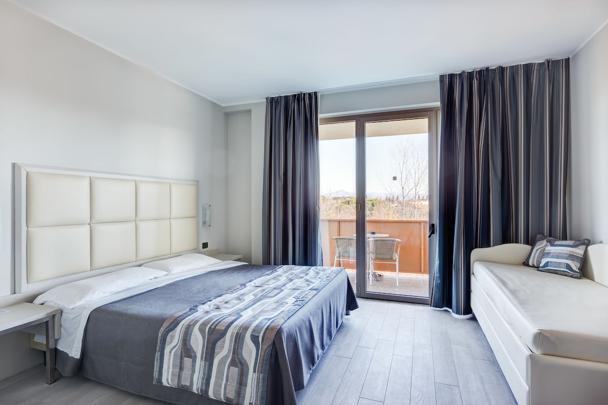 A hotel room with a bed, a sofa, a desk, and a sliding glass door leading to an outdoor area.