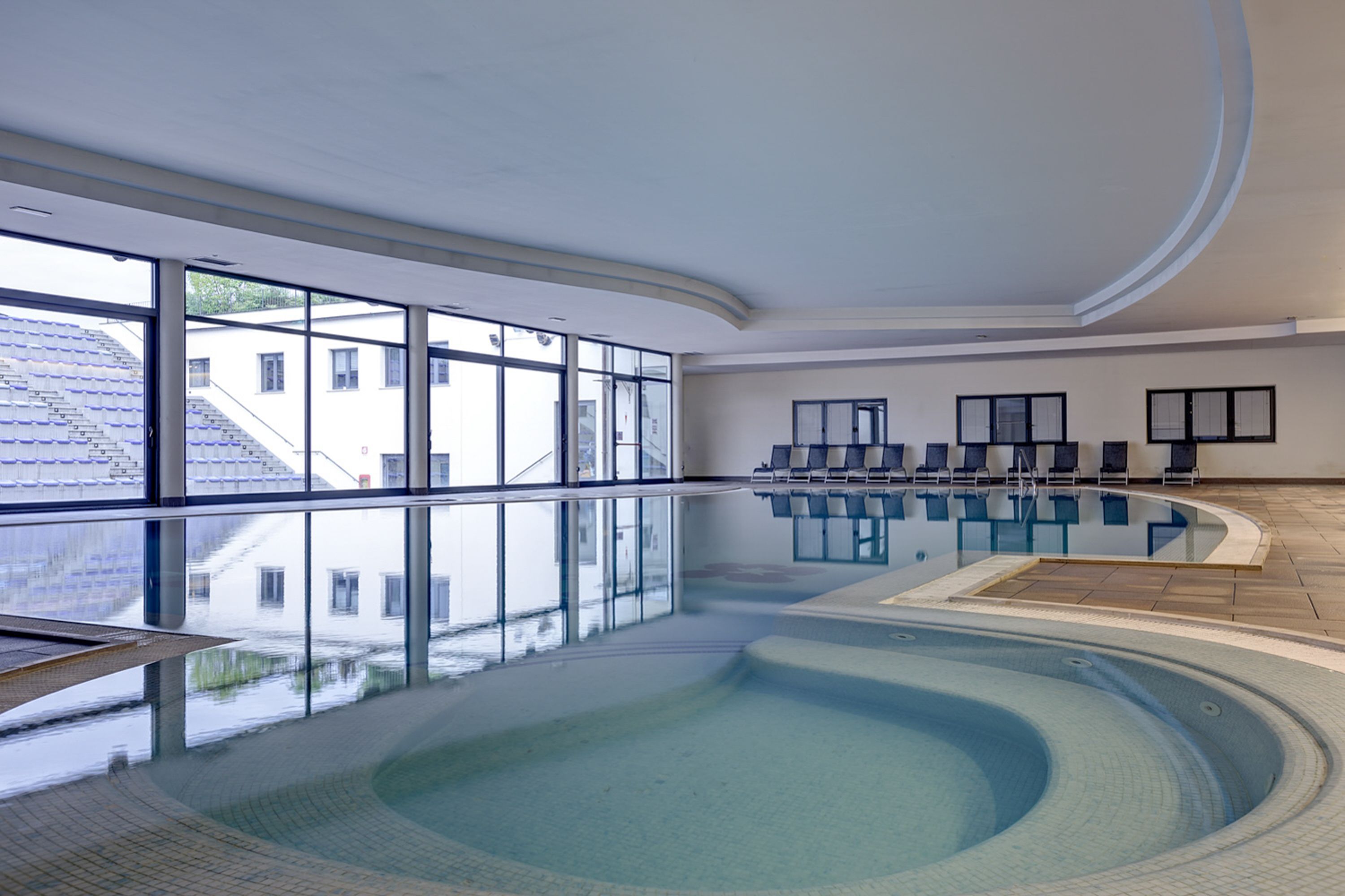A large indoor swimming pool with clear glass walls, a few lounge chairs, and a view of buildings outside.