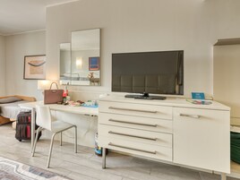 Habitación de hotel con un escritorio blanco, un televisor de pantalla plana, un espejo, una cama, una silla y una maleta.