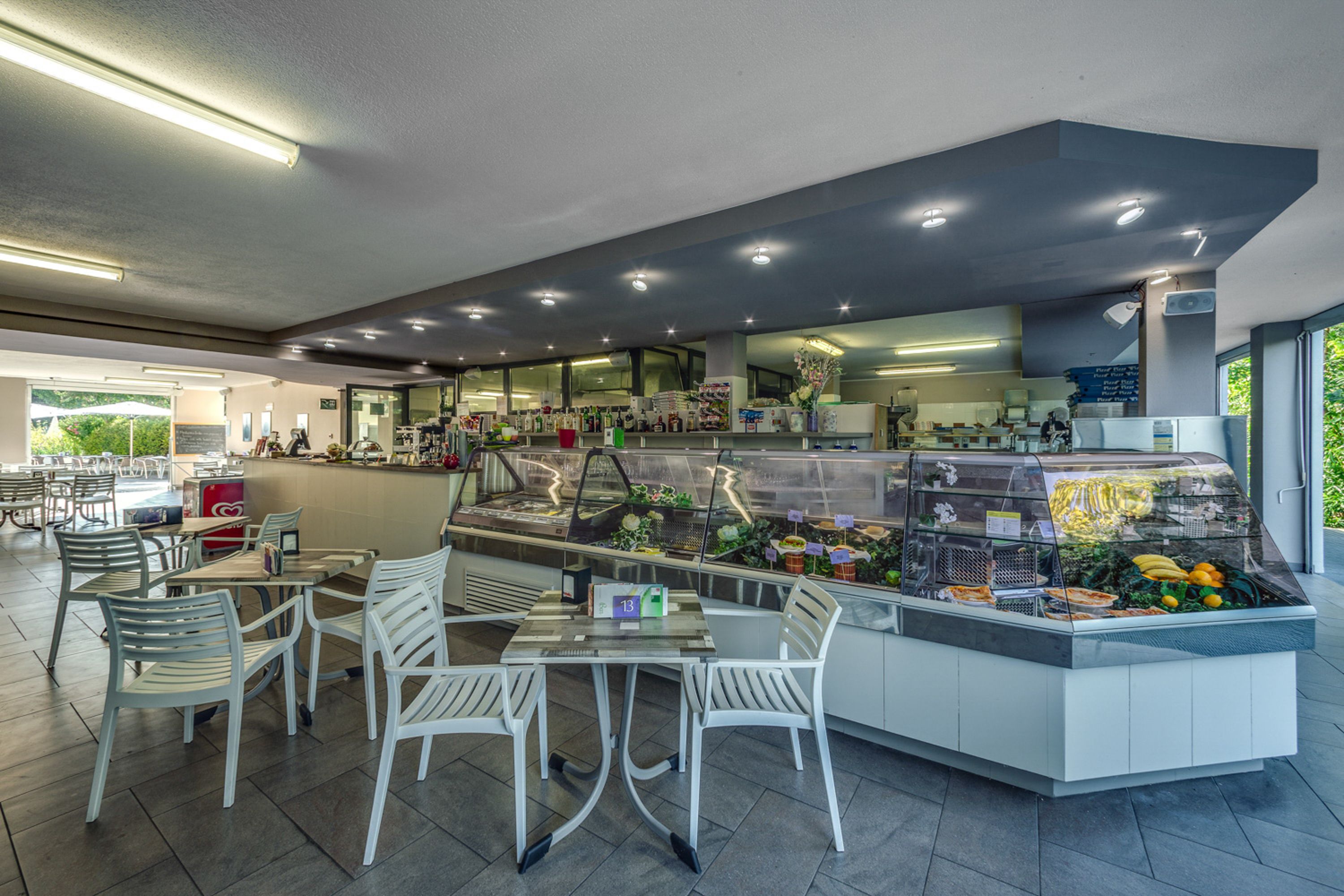 A modern cafe with a counter displaying fresh fruits and pastries, white chairs, and a clean, well-lit interior.