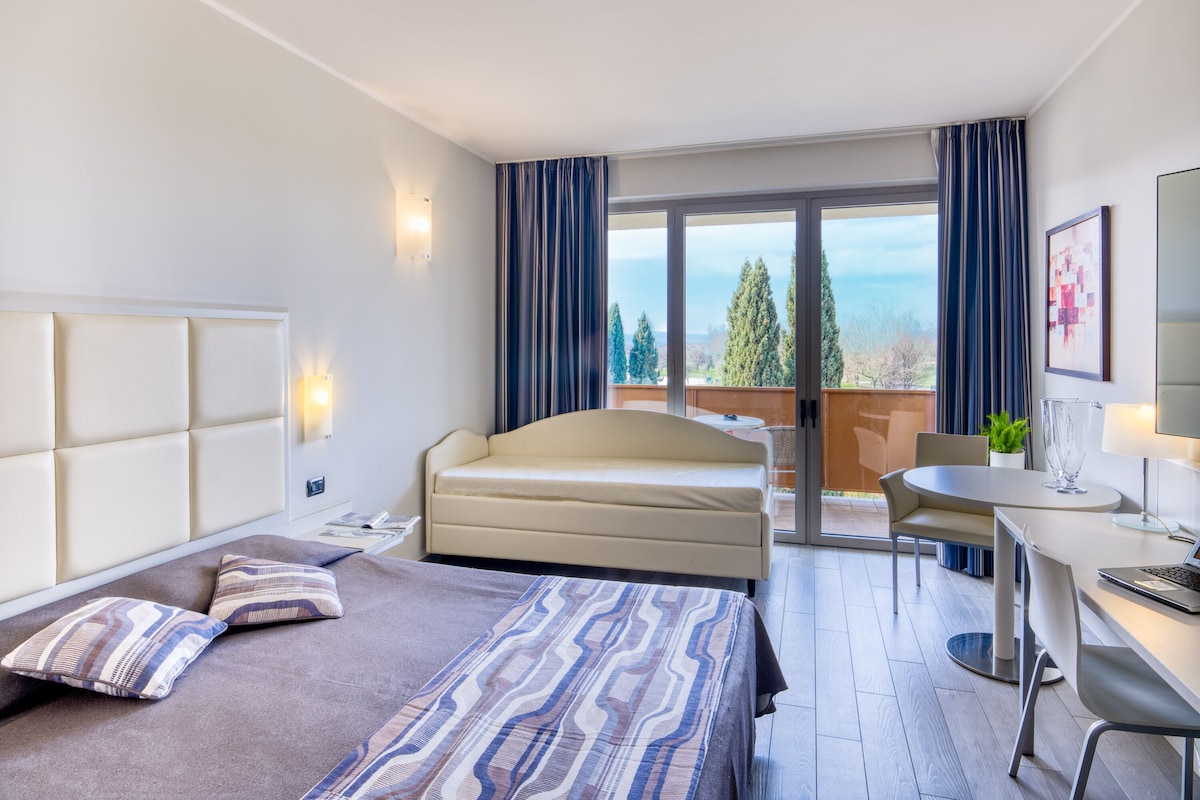 A modern hotel room with a bed, a desk, and a balcony with a view.