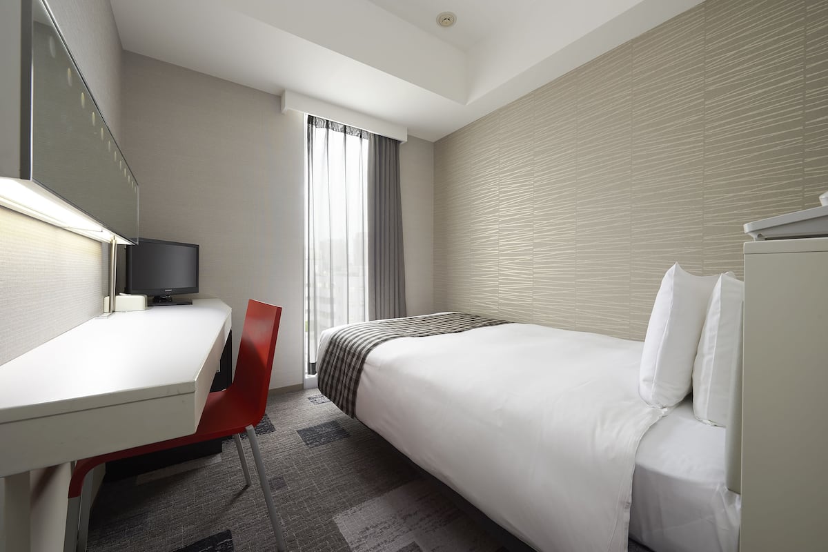 Une chambre d’hôtel comprenant un lit, un bureau avec une chaise, une télévision et une fenêtre avec des rideaux.