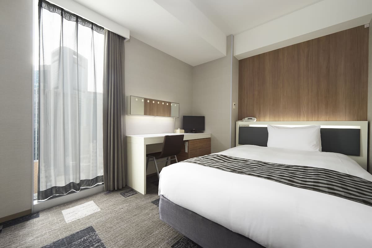 Une chambre d’hôtel avec un grand lit, un bureau avec une chaise, une télévision et une fenêtre avec des rideaux légers.