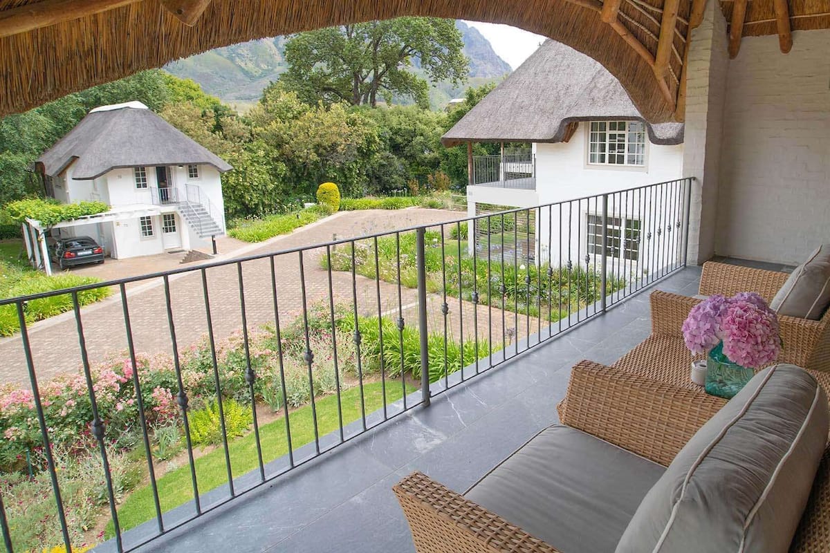 A balcony with wicker furniture and a view of a garden and two houses.