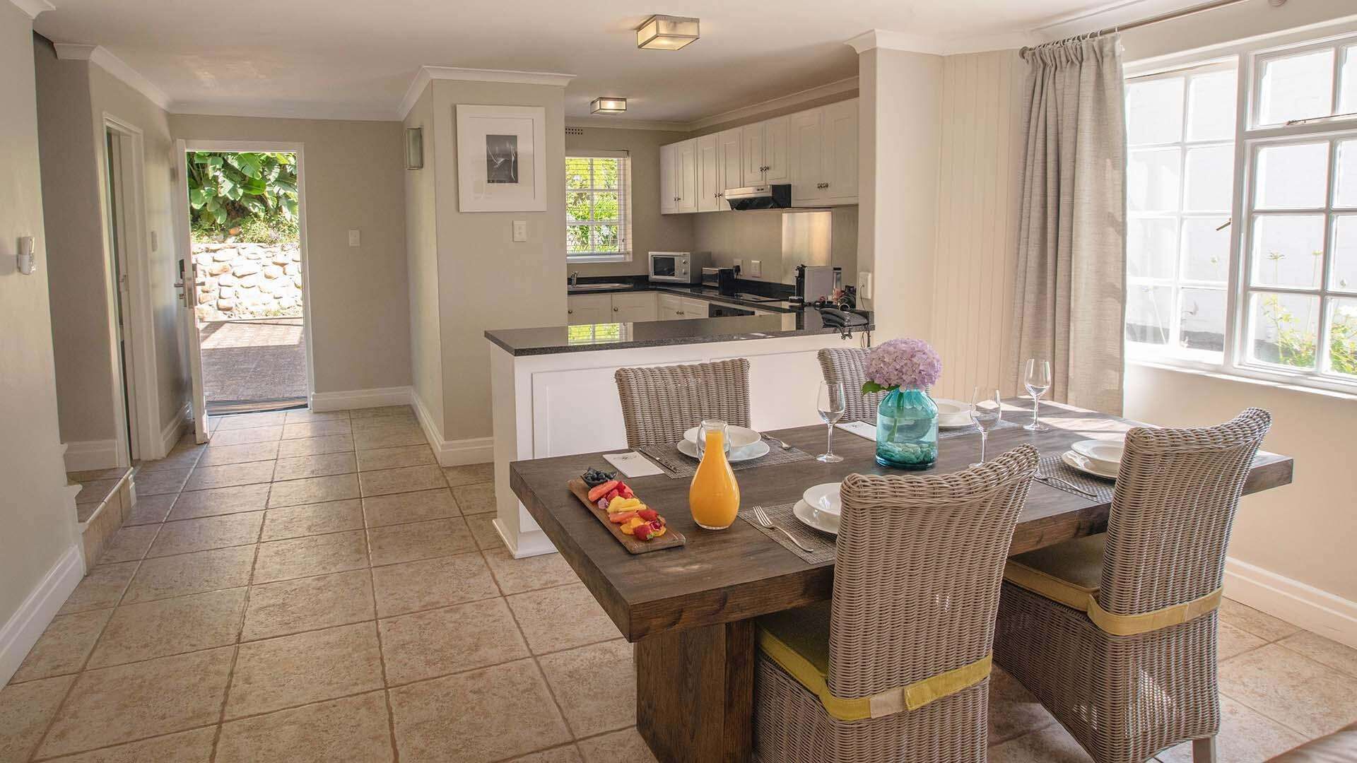 A dining area with a wooden table, wicker chairs, a fruit platter, a pitcher of orange juice, and a vase with flowers.