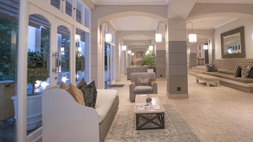 A spacious lobby with a sofa, armchairs, a coffee table, and a bench. There are large windows and a mirror on the wall.
