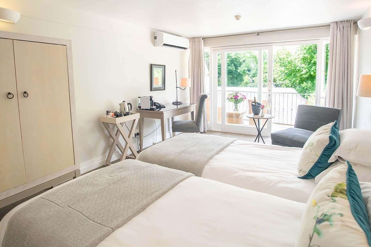 A hotel room with two beds, a desk, a chair, and a balcony with a view of greenery.