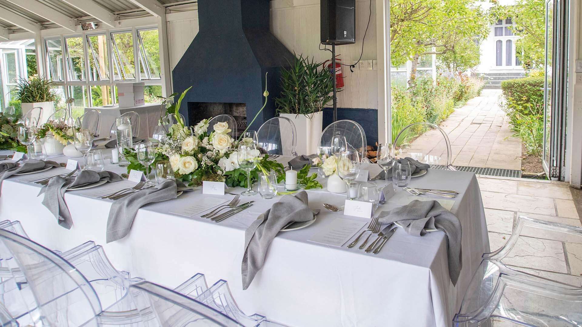 Une longue table à manger, dressée pour un événement formel, avec des chaises en verre transparent, des nappes blanches et des centres de table floraux.