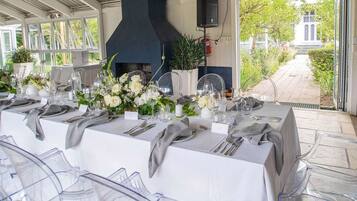 Une longue table à manger, dressée pour un événement formel, avec des chaises en verre transparent, des nappes blanches et des centres de table floraux.