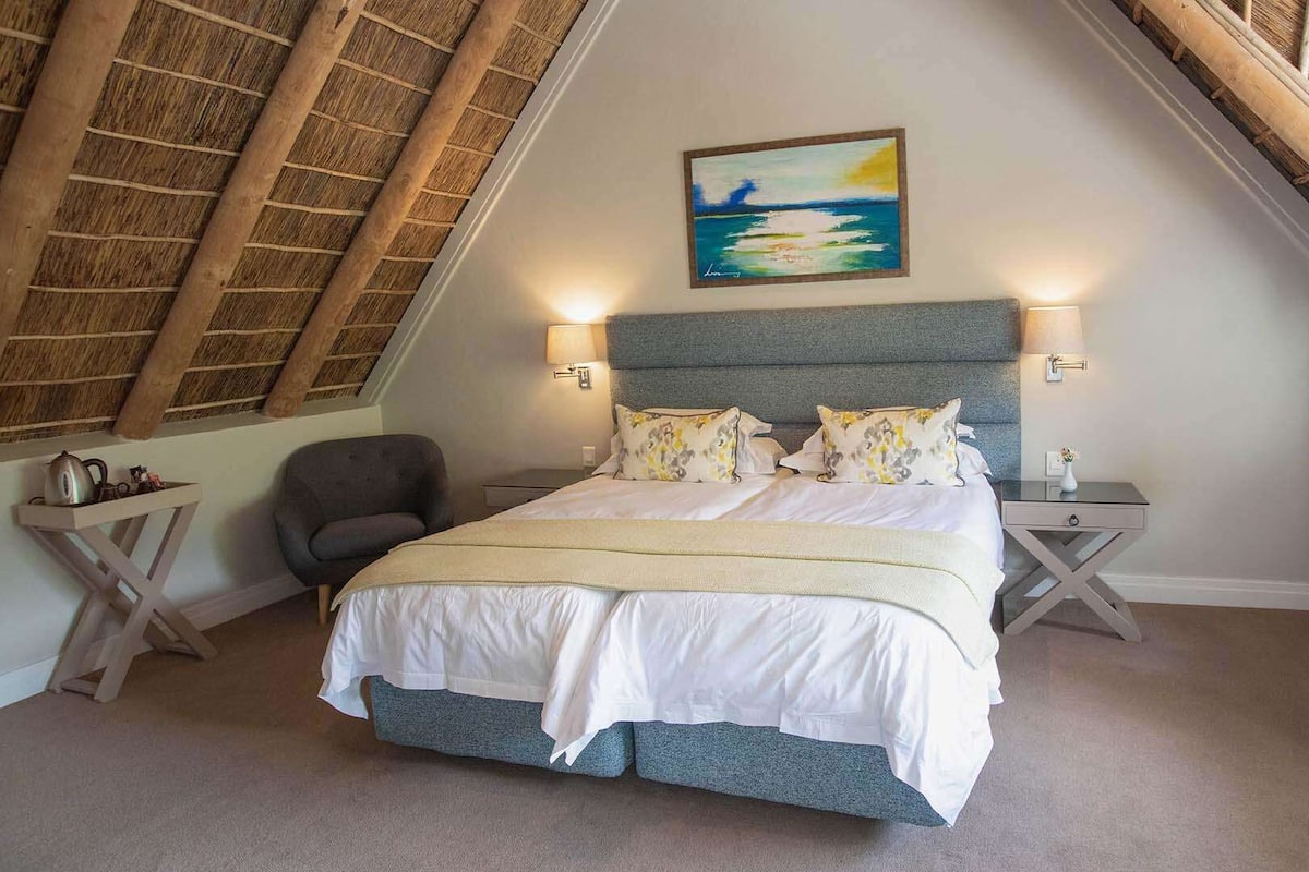 A bedroom with a sloped ceiling, a bed with white bedding, two bedside tables, a chair, and a painting on the wall.