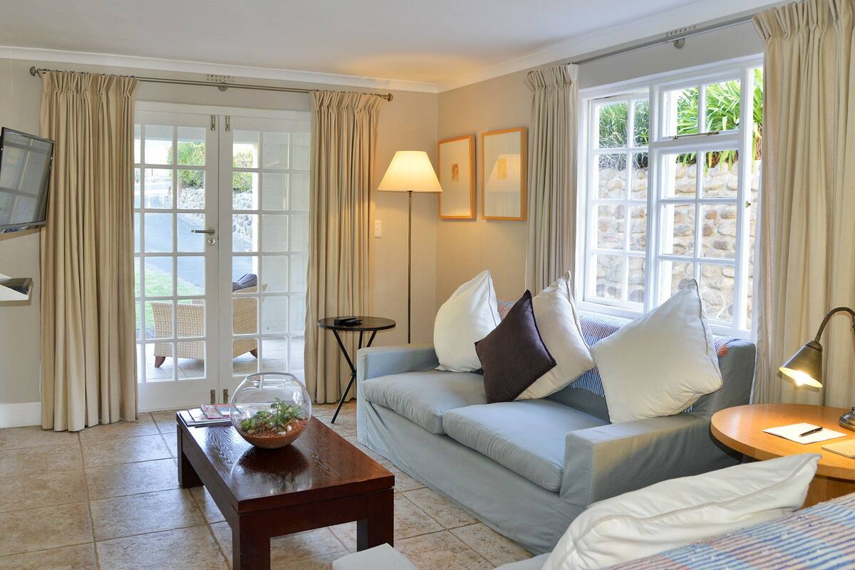 A cozy living room with a sofa, a coffee table, and a lamp. There is a TV mounted on the wall and a window with curtains.