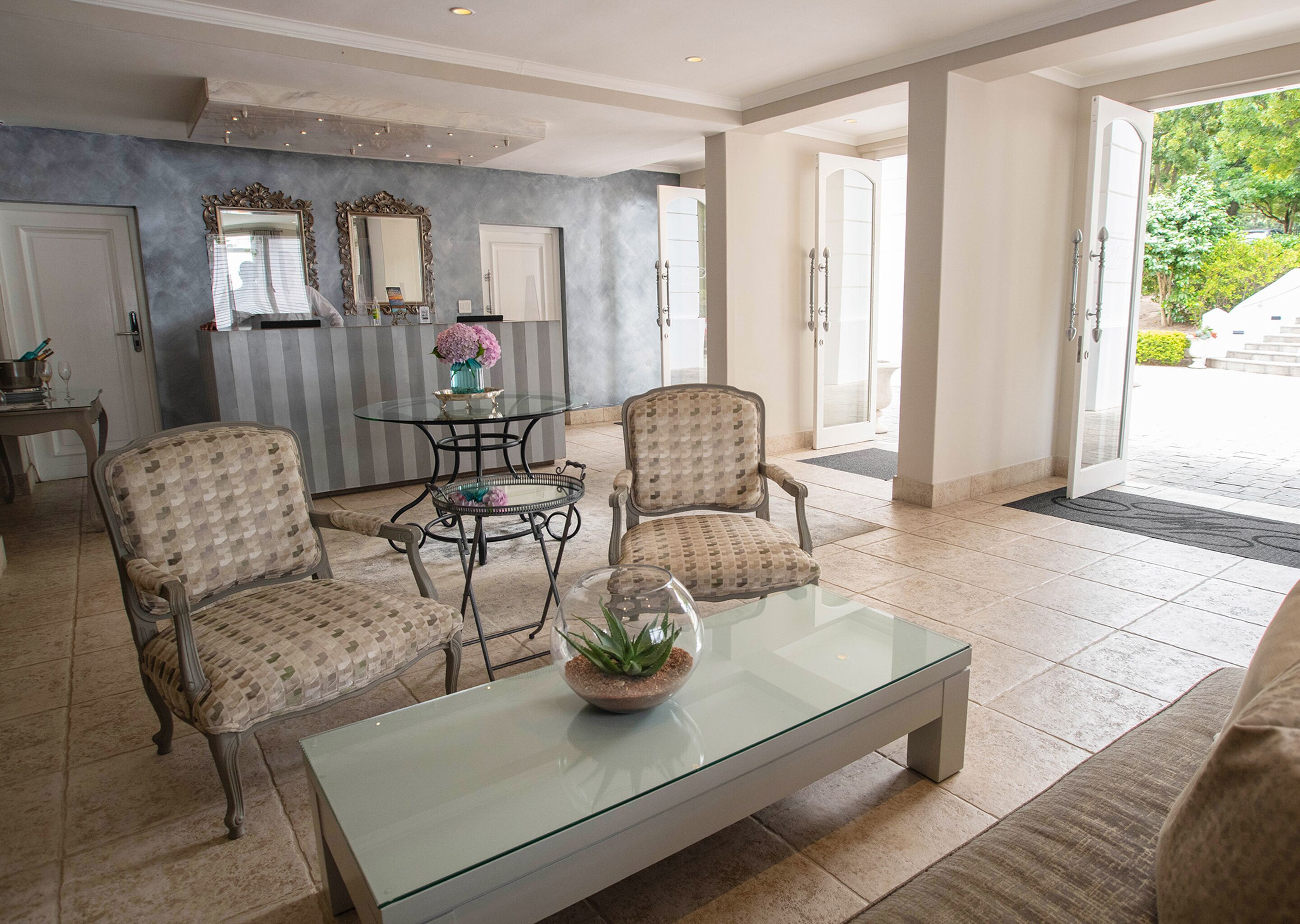 A spacious living area with a glass coffee table, two patterned armchairs, a small round table with a vase, and a large mirror.