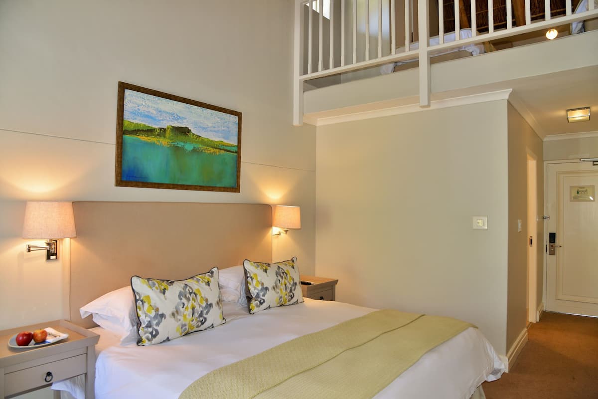 A hotel room with a bed, two bedside lamps, a nightstand with a plate of fruit, a painting on the wall, and a door to the right.