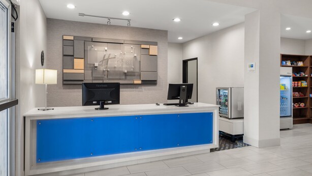 Un espace d’accueil d’hôtel avec un comptoir bleu, deux écrans, un réfrigérateur et un distributeur automatique.