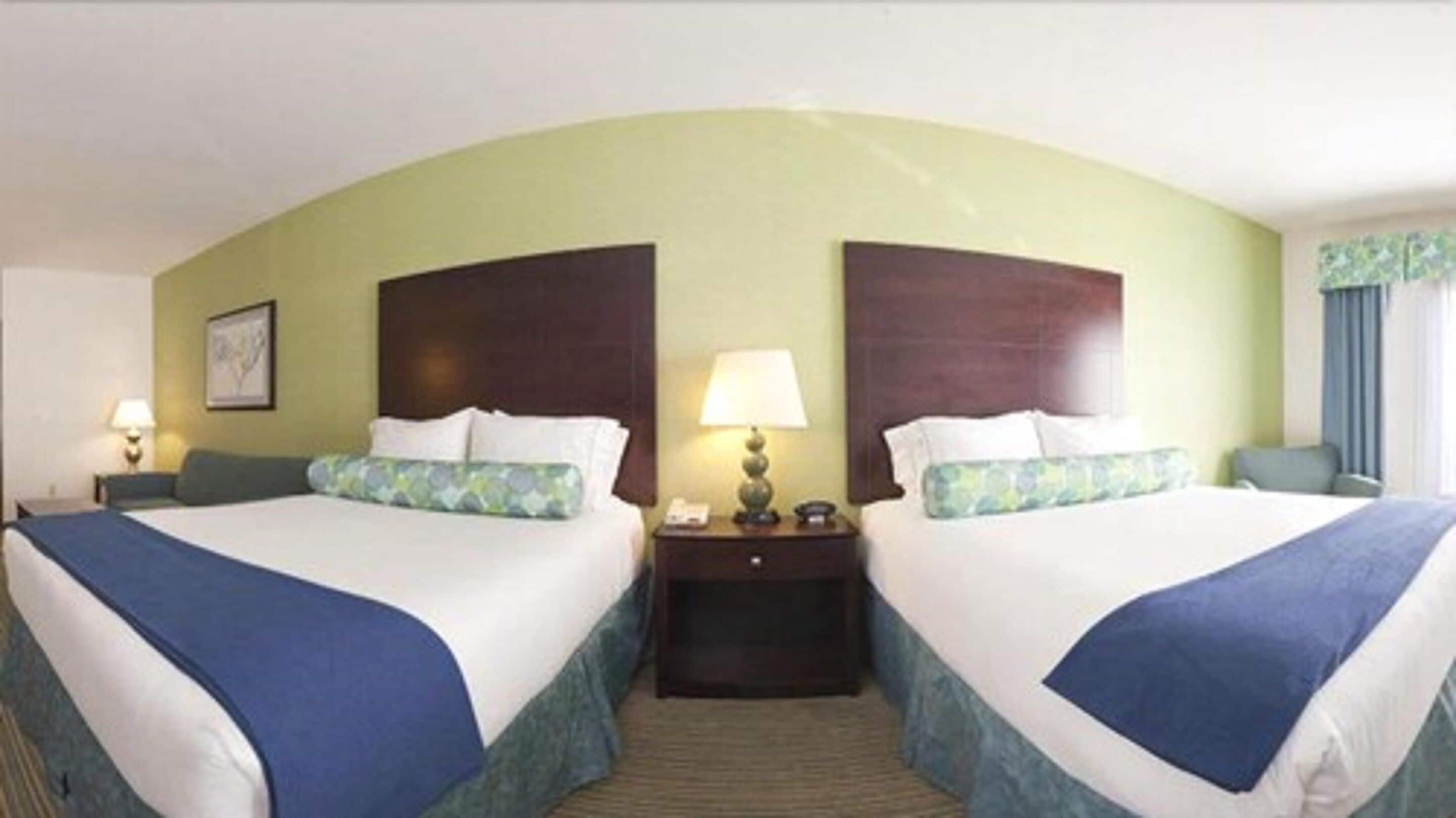 A hotel room with two beds, each with a blue bedspread and matching pillows, a nightstand with a lamp, and a framed picture on the wall.