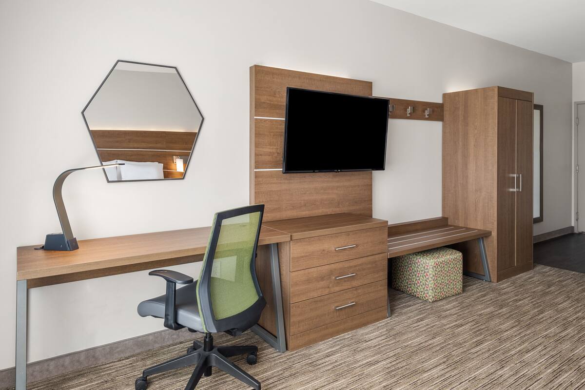 Ein modernes Hotelzimmer mit Schreibtisch aus Holz, einem Flachbildfernseher, einem grünen Bürostuhl, einem Kleiderschrank und einem gemusterten Teppich.