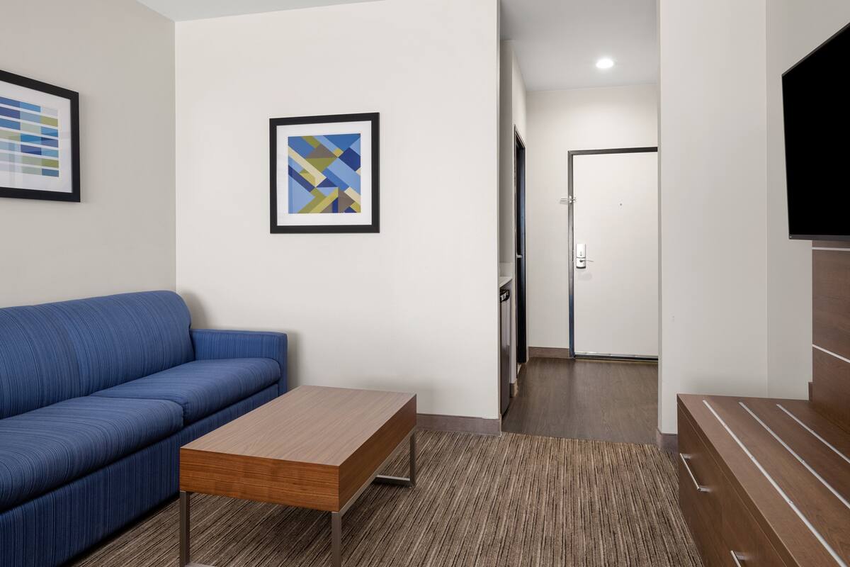 Ein modernes Hotelzimmer mit einem blauen Sofa, einem Holztisch, einem gerahmten abstrakten Kunstwerk und einem an der Wand befestigten Flachbildfernseher.