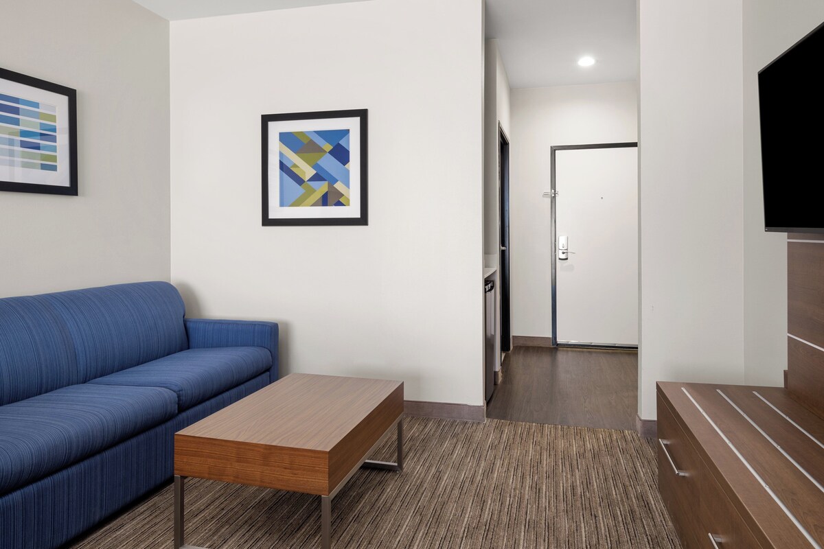 A modern hotel room with a blue sofa, a wooden coffee table, a framed abstract art piece, and a flat-screen TV mounted on the wall.
