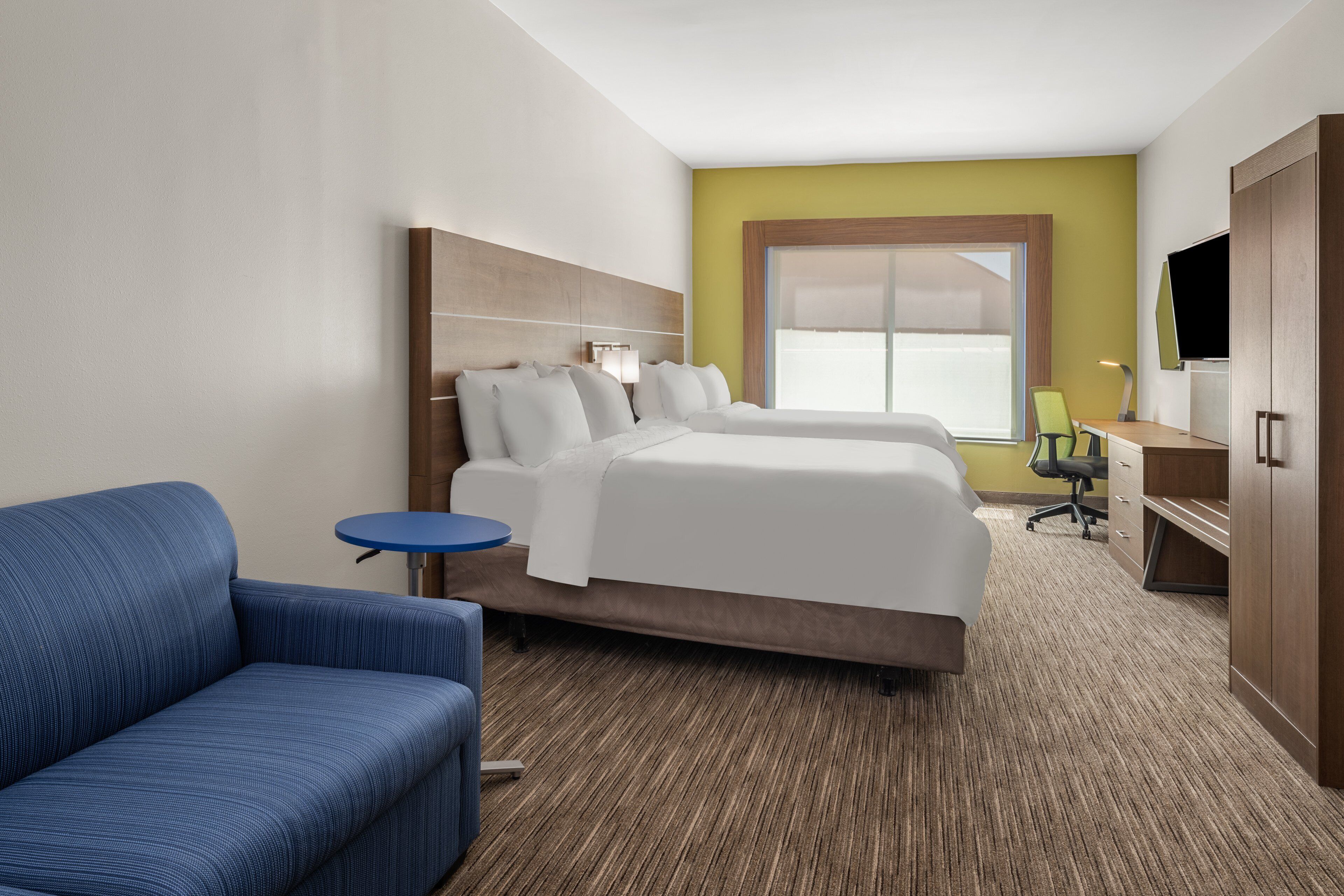 A hotel room with a large bed, a blue armchair, a small round table, a desk with a chair, and a TV on a wooden cabinet.
