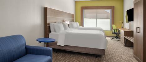 A hotel room with a large bed, a blue armchair, a small round table, a desk with a chair, and a TV on a wooden cabinet.
