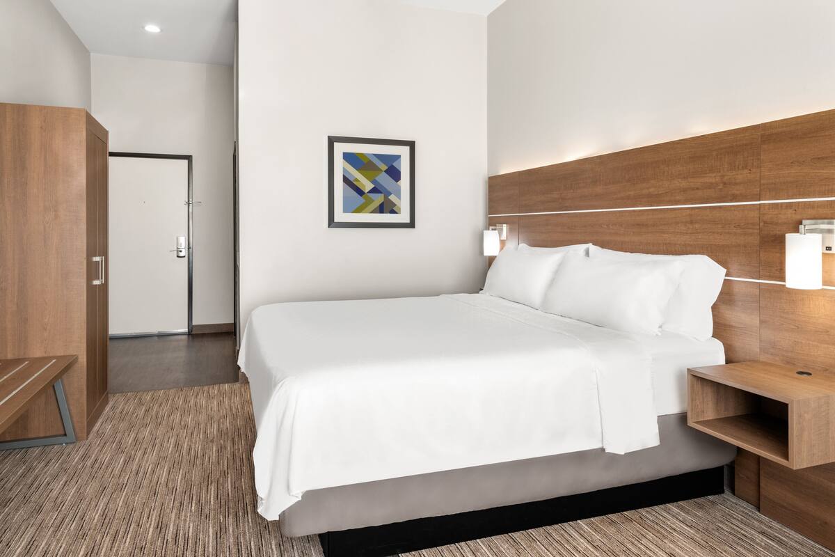 Ein Hotelzimmer mit einem großen Bett, einem Holz-Kopfteil, Nachttischen mit Lampen, einem gerahmten abstrakten Kunstwerk an der Wand und einer Holzbank.