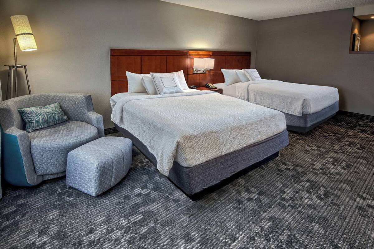 A hotel room with two beds, a chair, a lamp, and a carpeted floor.