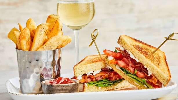 A sandwich cut in half, a glass of white wine, and a metal container of fries.